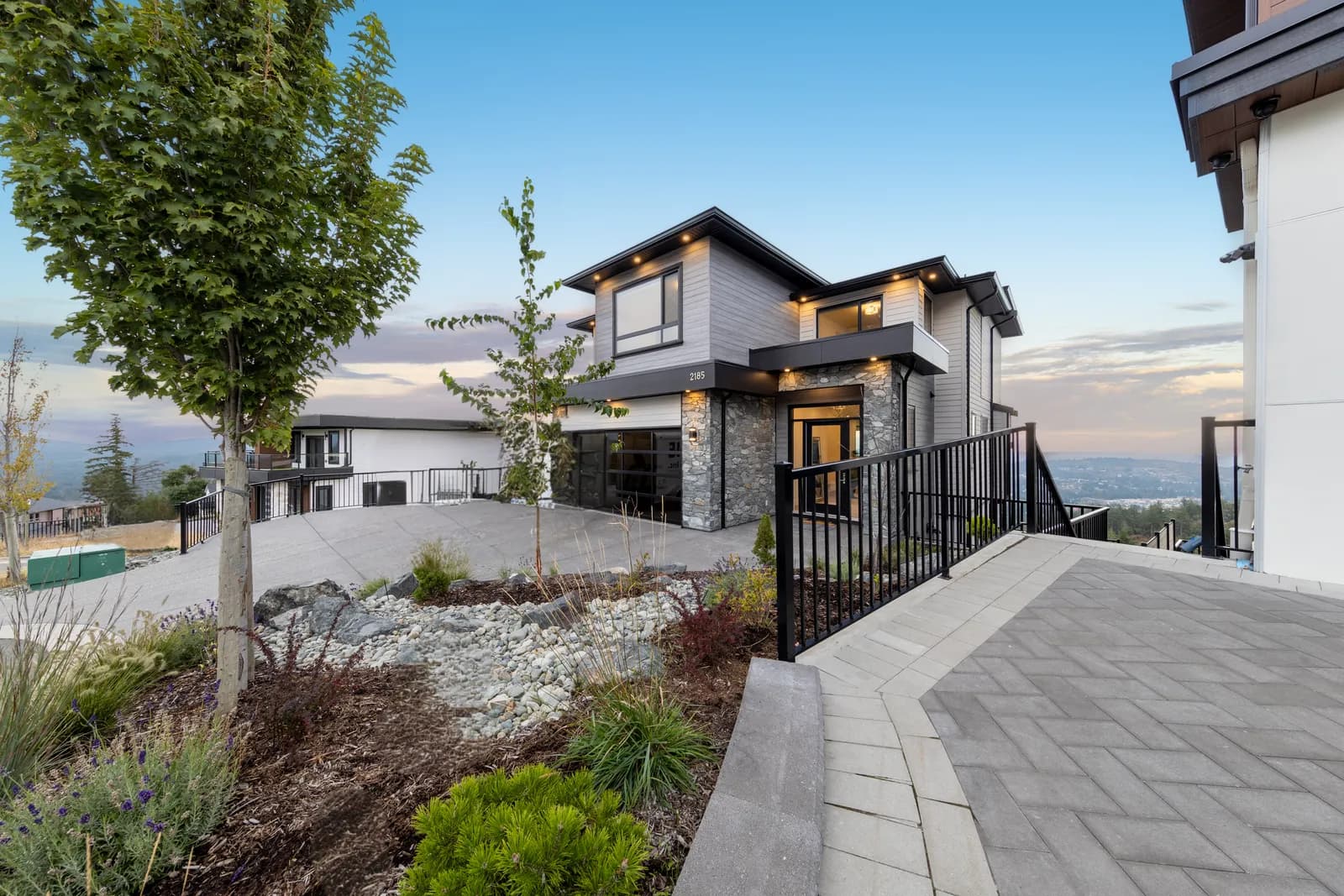 Exterior of the custom new home at 2185 Navigators Rise, Bear Mountain, Langford BC at dusk — stone and board-and-batten facade, aggregate driveway and hillside views over Langford, built by Pro Construction Group